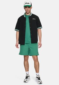 Zwarte blouse met groene accenten, groene t-shirt eronder, groene shorts en zwart/witte sneakers. FILA-logo op het shirt en de pet.