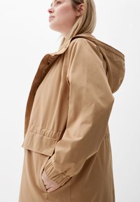 Beige Kapuzenjacke mit lockerer Passform, ausgestattet mit elastischen Bündchen und einem Druckknopfverschluss auf der Vorderseite. Glattes Material mit schlichtem Design und seitlichen Taschen.