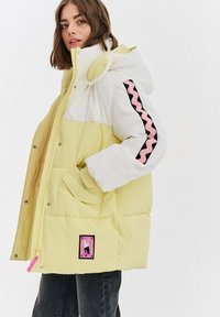 Prešívaná bunda s dizajnom farebného bloku v žltej a bielej farbe, s ružovým vlnkovým pruhom na čiernom rukáve a logom na nášivke.