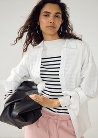 Femme aux cheveux bouclés portant de grandes boucles d'oreilles créoles, chemise blanche par-dessus un pull rayé, pantalon rose, tenant un sac en cuir noir sur un fond clair.