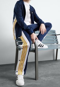 Chaqueta de chándal azul marino y pantalones con rayas amarillas y blancas, combinados con zapatillas blancas con rayas negras. Tejido suave, corte relajado.