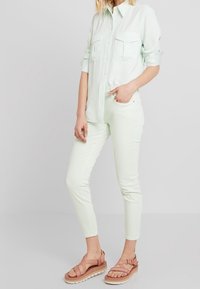 Persona vestida con una camisa de botones verde claro con bolsillos en el pecho, pantalones slim a juego y sandalias de plataforma rosas con tiras.