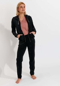 Tuta in velluto nero con cappuccio zip e pantaloni affusolati, abbinato a una maglietta a V rosa chiaro con taschino. Texture morbida.