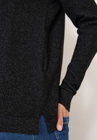 Czarny dzianinowy sweter o teksturowanym wykończeniu, z prążkowanymi mankietami i bocznymi rozcięciami u dołu. Noszony z niebieskimi dżinsami.