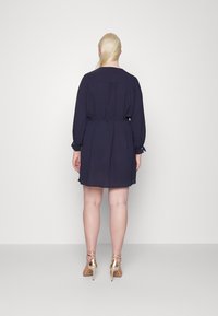 Vestido curto azul marinho com mangas longas, cintura cingida por um cinto e punhos com botões, apresentando uma textura fluida e pregas sutis nas costas.