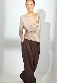 Haut beige drapé à manches longues avec un décolleté profond, associé à un pantalon brun à taille haute et plis, ample. Texture de tissu lisse.