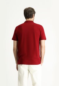 Polo shirt rosso a maniche corte realizzato in tessuto liscio, con colletto classico e design aderente, mostrato su una persona vista da dietro.