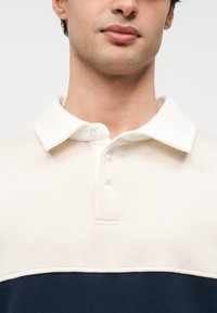 Πόλο πουκάμισο με κρεμ επάνω μέρος και ναυτικό μπλε κάτω μέρος, κατασκευασμένο από απαλό ύφασμα, με κλασικό γιακά και εμπρόσθιο κλείσιμο με τρία κουμπιά.