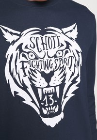 Sweat-shirt bleu marine avec un grand graphisme blanc représentant un visage de tigre, accompagné du texte "SCHOTT FIGHTING SPIRIT" et du chiffre "13" intégrés au design.