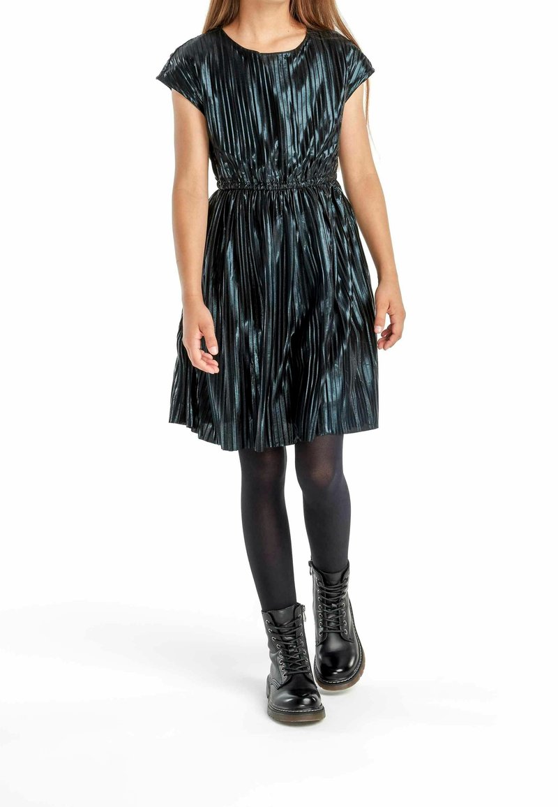 Fille portant une robe plissée noire brillante, des collants noirs et des bottes noires à lacets, marchant en avant sur un fond blanc.