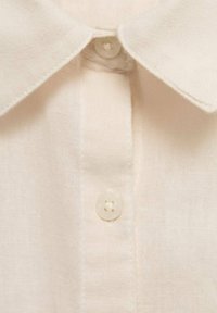 Camicia in lino beige con colletto e chiusura a un pulsante. La texture è morbida con dettagli di cucitura visibili.