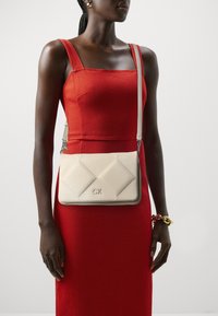 Borsa a tracolla beige trapuntata con accessori in argento, che presenta il logo "CK". Abbinata a un abito rosso aderente. Tracolla regolabile.