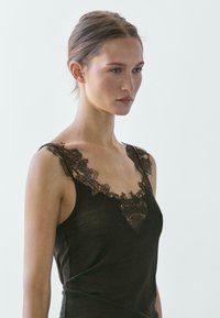 Giovane donna con capelli raccolti che indossa una canottiera nera con rifiniture in pizzo sullo scollo e sulle spalle, rivolta verso destra.