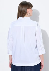 Camicia bianca a maniche lunghe con polsini abbottonati e una piega sul retro. Tessuto leggero con una texture liscia, caratterizzato da una leggera lucentezza.