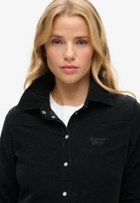 Sort fløjljakke med høj krave, der har trykknapper og "Superdry"-logo broderet i mørk tråd på brystet.
