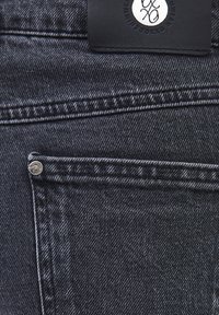 Vaqueros de mezclilla negros con un tejido texturizado, que cuentan con un pequeño bolsillo frontal, herrajes plateados y una etiqueta de cuero negro en la cintura.