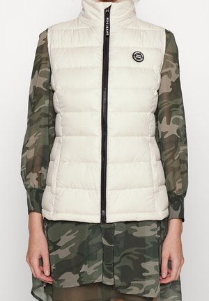 Biała pikowana kamizelka bez rękawów z czarnym zamkiem i naszywką z logo, noszona na długiej, zielonej sukience w kamuflażu.