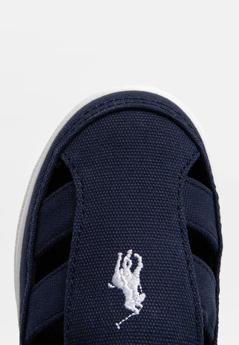 Chaussure à enfiler en tissu navy avec semelle en caoutchouc blanche, présentant un logo brodé distinctif. Design ouvert, surface texturée et sangles sécurisées.