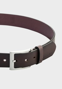 Brązowy skórzany pasek z metalową klamrą w kolorze srebrnym, o gładkiej fakturze i kilku otworach do regulacji. Minimalistyczny design.