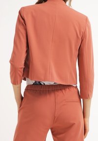 Krótka marynarka z tkaniny w odcieniu przydymionego różu, z rękawami do łokcia, posiadająca gładki szew z tyłu oraz gładką, fakturowaną powierzchnię.