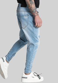 Svijetloplavi traper traperice u stilu joggera s uskim krojem, s bočnim patentnim zatvaračima, stražnjim džepovima i suptilnim detaljima izbjeljivanja. U kombinaciji s bijelim tenisicama.