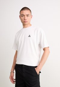 Man with short hair wearing a white ACG logo T-shirt and black pants, standing with one hand in pocket against a plain background.