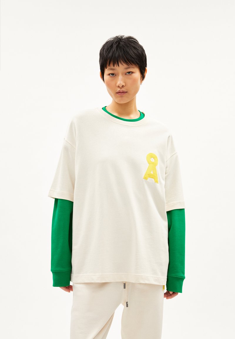 Jong iemand met kort zwart haar die een gelaagde outfit draagt met een witte oversized T-shirt en groene lange mouwen eronder.