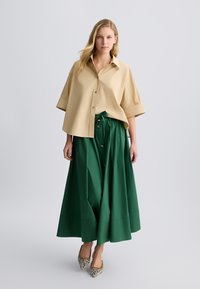 Camicia beige a maniche corte con bottoni abbinata a una lunga gonna verde fluida con cintura e bottoni dorati in evidenza; scarpe con motivo di pelle di serpente.