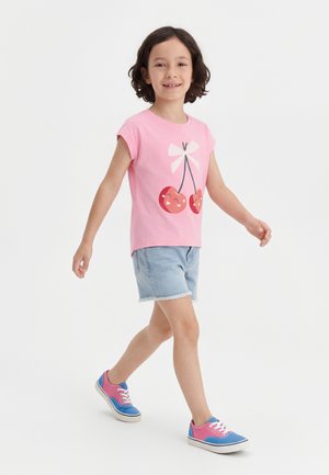 Jeune enfant marchant avec un t-shirt rose à motif cerises, un short en jean et des baskets colorées rose et bleue sur fond blanc.