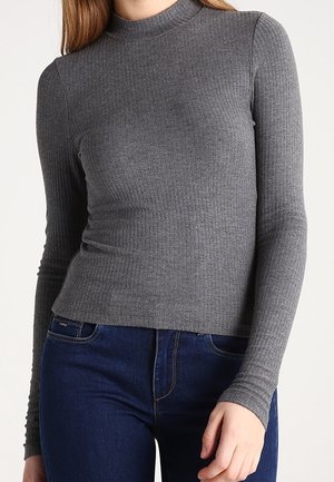 Vrouw draagt een aansluitend geribd grijs topje met lange mouwen en een col, gecombineerd met donkerblauwe jeans, tegen een effen achtergrond.