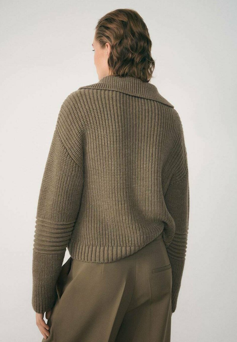 Jersey de punto en verde oliva con una textura acanalada, corte oversize, cuello ancho y puños enrollados, combinado con pantalones de traje.