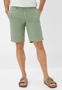 Pantaloni corti verde oliva con tasche, realizzati in tessuto leggero. Presentano una silhouette dritta e orli cuciti. Abbinati a sandali marroni.