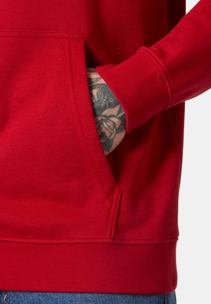 Mano con tatuaje floral parcialmente dentro del bolsillo frontal de una sudadera roja, combinada con jeans azules.