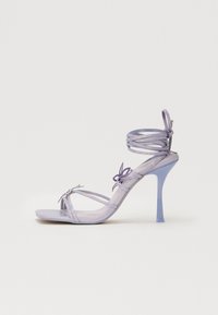 Sandale à talons hauts violette avec des brides transparentes, détail de nœud décoratif et talon aiguille fin. Texture lisse et design épuré.