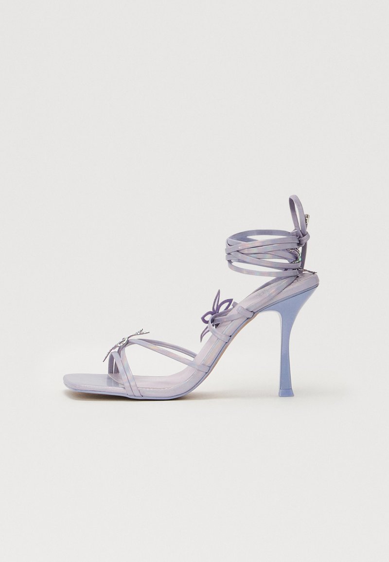 Sandale à talons hauts violette avec des brides transparentes, détail de nœud décoratif et talon aiguille fin. Texture lisse et design épuré.