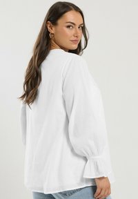 White long-sleeve blouse with puffed sleeves and gathered cuffs, featuring a round neckline and a relaxed fit, paired with light blue jeans.