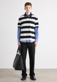 En svart og hvit stripet polo skjorte over en lyseblå knappskjorte, sammen med svarte bukser og en svart lær tote bag. Jevn tekstur.