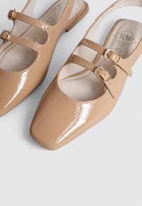 Ballerines en cuir verni beige avec un bout carré. Deux brides réglables avec des boucles dorées sur le cou-de-pied. Fini lisse et brillant.
