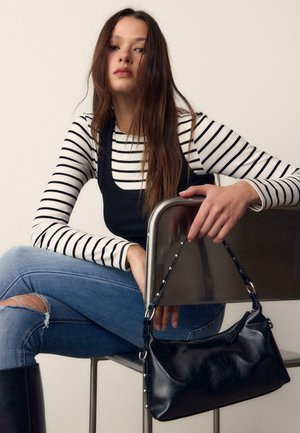 Young woman with long hair wearing striped shirt and black tank top, ripped blue jeans, sitting on chair holding black leather handbag.