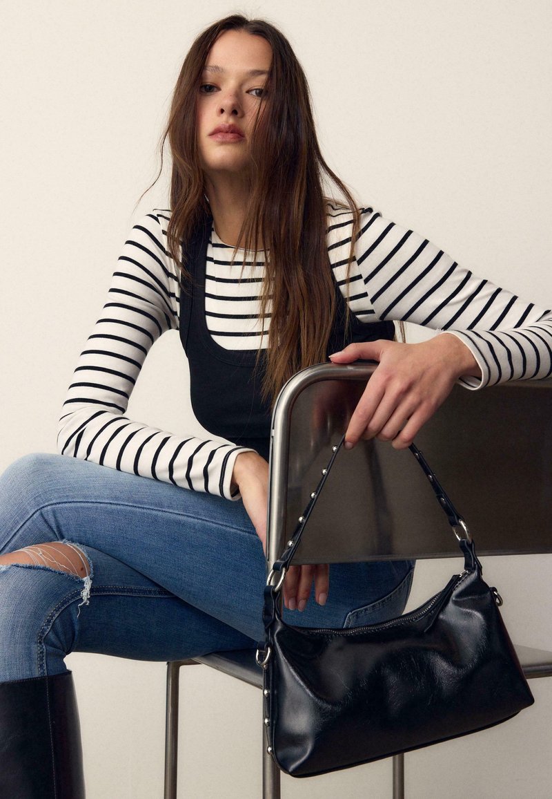Giovane donna con capelli lunghi che indossa una maglietta a righe e un top nero, jeans blu strappati, seduta su una sedia con una borsa di pelle nera in mano.