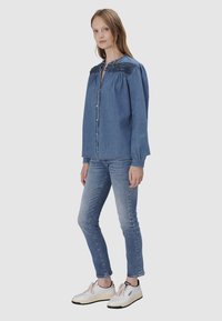 Koszula dżinsowa z haftowanymi niebieskimi detalami na kołnierzyku, z zapięciem na guziki i długimi rękawami. Połączona z jasnoniebieskimi obcisłymi spodniami i białymi trampkami.