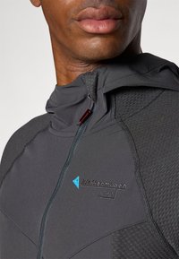 Veste de sport grise avec fermeture éclair à l'avant, capuche, accents texturés et logo brodé en bleu. Tissu lisse et respirant.