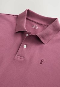 Polo tričko s krátkym rukávom v odtieni slivkovej, vyrobené z textúrovanej bavlny. Obsahuje rebrovaný golier, tri biele gombíky a malý vyšívaný detail.