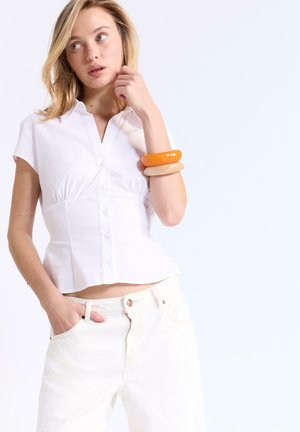 Blonde woman wearing a fitted white short-sleeve blouse and white pants, accessorized with two chunky bracelets on her wrist.