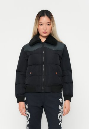 Woman with long ombre hair wearing a black puffer jacket with a shearling collar and black pants with white floral embroidery.