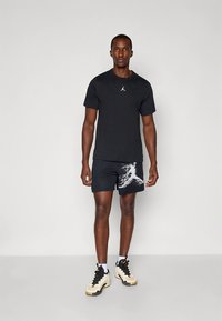 Svart t-shirt med kort ärm och vit logotyp, kombinerad med mönstrade svarta shorts och elfenbensvita sneakers med svarta detaljer. Enkelt design.