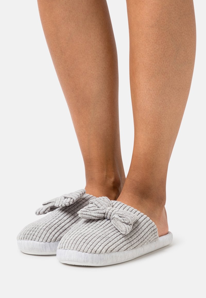 Chaussons en velours côtelé gris avec tissu texturé, ornés d'un détail nœud sur le dessus et une doublure blanche douce, style ouvert à l'arrière.