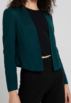 Vrouw draagt een donkergroene blazer met open voorkant over een zwart cropped topje en zwarte broek, getoond van nek tot heupen.