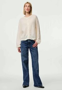 Cardigan léger beige, transparent, avec un devant à boutons ; associé à un jean bleu foncé à jambes larges. Texture lisse, coupe ample. Chaussures noires.
