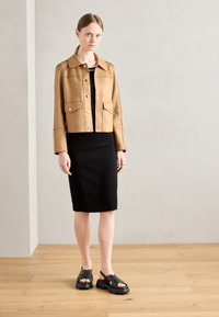 Ruskea nubukista valmistettu lyhyempi takki, jossa on kaksi etutaskua, yhdistetty mustaan tiukkaan mekkoon ja mustiin korkokenkiin, joissa on ristittyjä hihnoja.